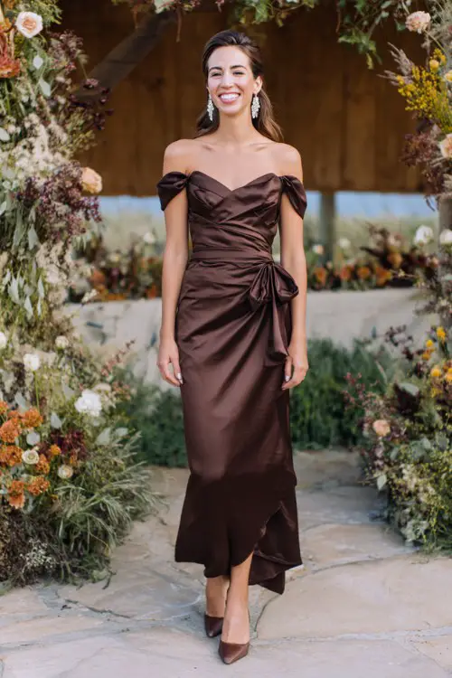 A woman wears a chocolate brown satin gown with an off-shoulder neckline and cascading side ruffle