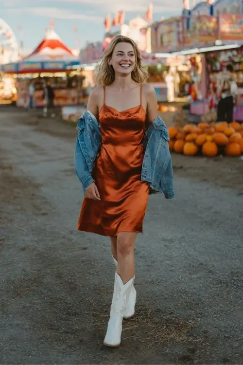 A woman wears a copper satin slip dress with a denim jacket and white cowboy boots outfit for a fall country concert