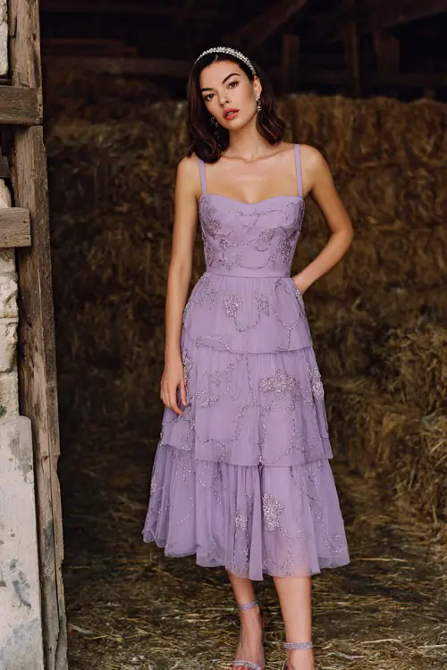 A woman wears a lilac tea-length dress with a fitted bodice and layered chiffon skirt embroidered with silver vines