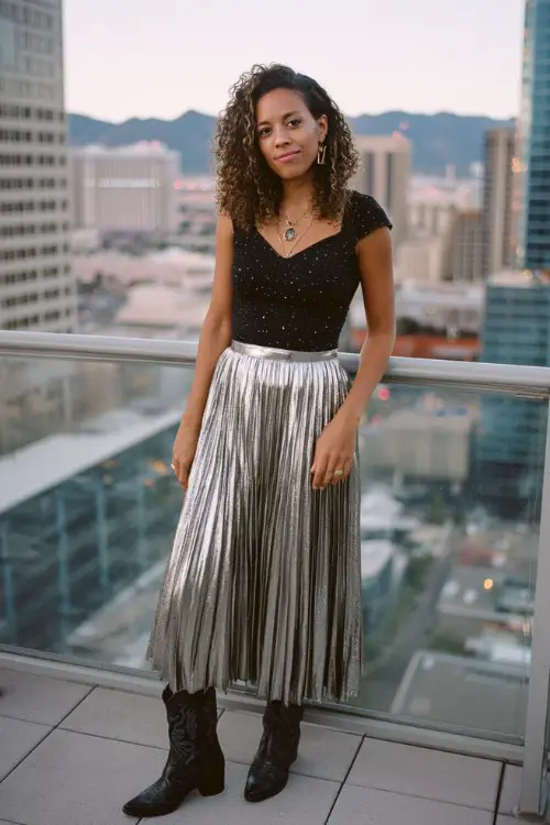 A woman wears a metallic pleated skirt with a fitted black top featuring subtle sparkle, paired with black cowboy boots