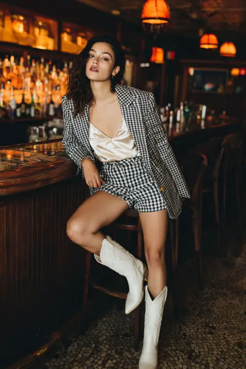 A woman wears a satin camisole, faux leather shorts, an oversized houndstooth blazer, and white cowboy boots