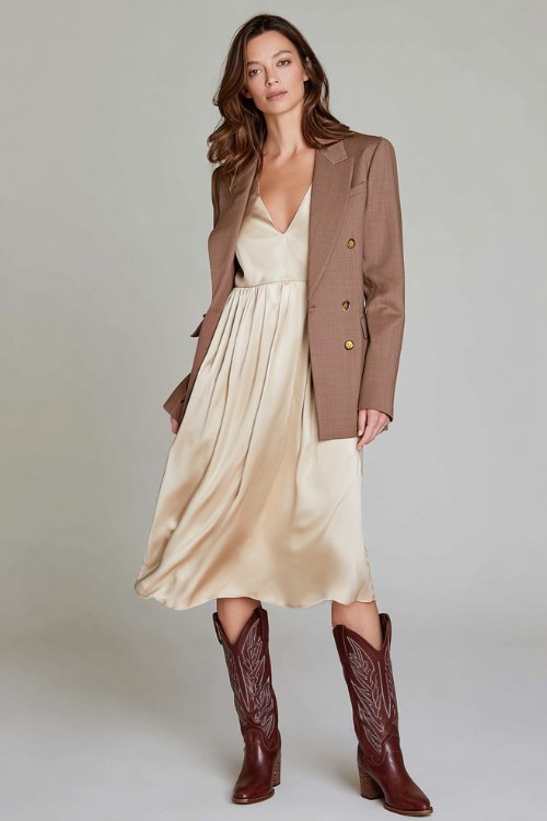 A woman wears a silky beige midi dress with a modest V-neckline and fluid movement, layered under a sharp double-breasted blazer and paired with tall brown cowboy boots