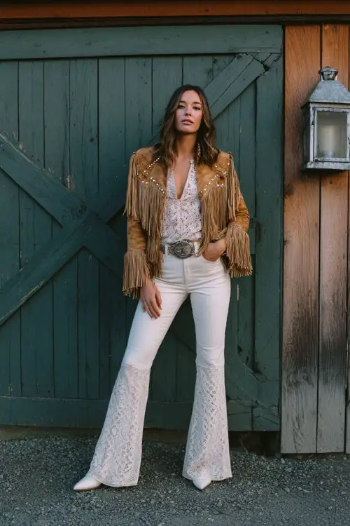 A woman wears flared jeans with a tucked-in ivory lace blouse, a suede fringe jacket, and white leather cowboy boots