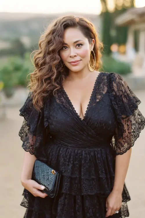 A curvy woman wears a black lace dress featuring ruffled lace sleeves, a wrap-style bodice, and tiered lace layers cascading through the skirt