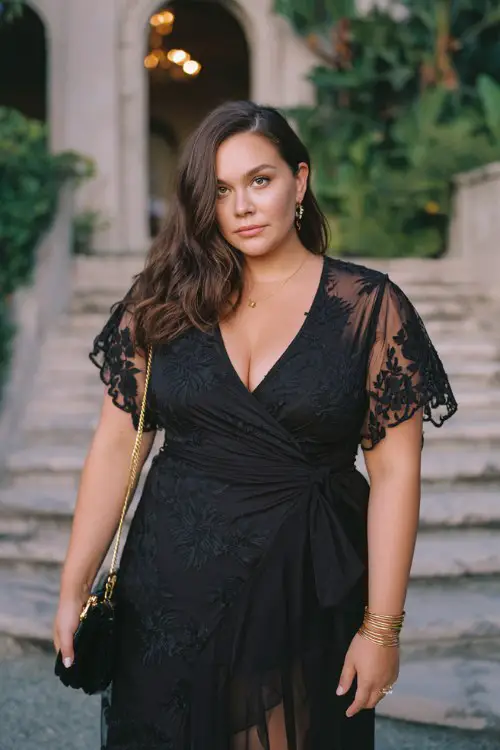 A curvy woman wears a black lace wrap dress with floral lace embroidery, flutter sleeves, and a flowing asymmetrical hemline