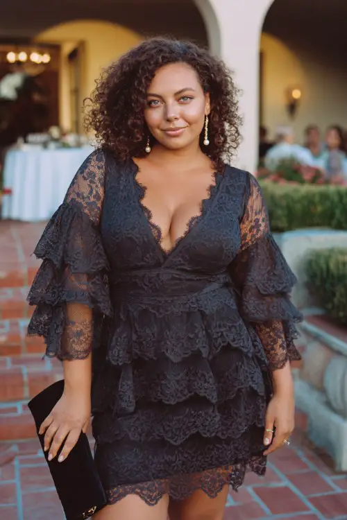 A curvy woman wears a dark lace tiered mini dress featuring flutter sleeves and delicate floral lace layers, styled with pearl drop earrings and a satin clutch