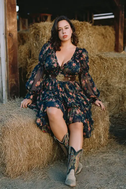 A curvy woman wears a navy floral chiffon dress with a tiered skirt and sheer sleeves, styled with embroidered cowboy boots, a cinched belt