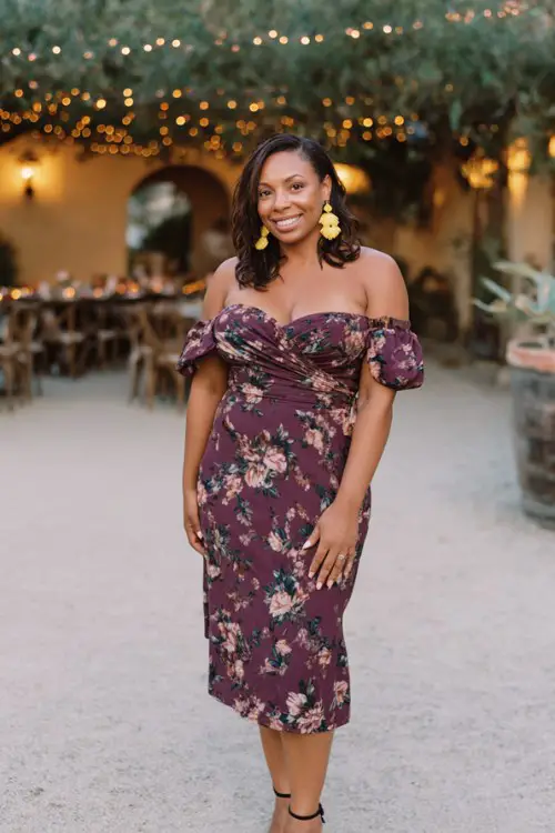 A curvy woman wears a plum floral off-shoulder midi dress with a fitted waist, paired with heeled sandals and bold statement earrings
