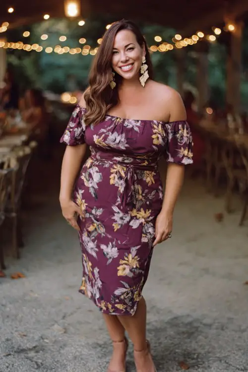 A curvy woman wears a plum floral off-shoulder midi dress with a fitted waist, paired with heeled sandals and bold statement earrings