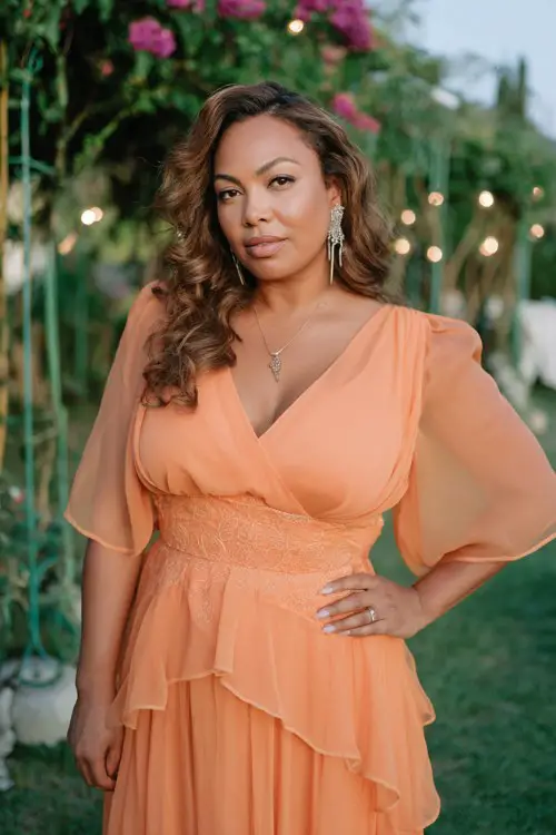 A curvy woman wears a sunset-orange chiffon dress featuring puff sleeves, embroidered waist details, and cascading ruffle tiers along the skirt