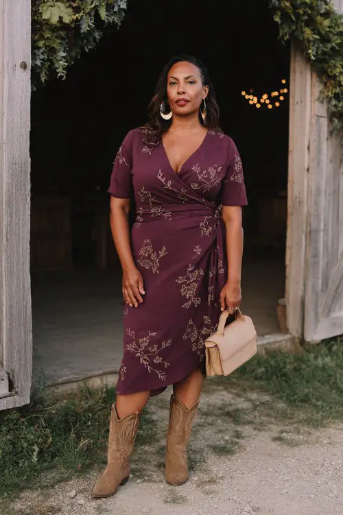A curvy woman wears an elegant wrap midi dress in deep plum with embroidered floral vines and a defined waist, styled with suede cowboy boots, a structured mini bag