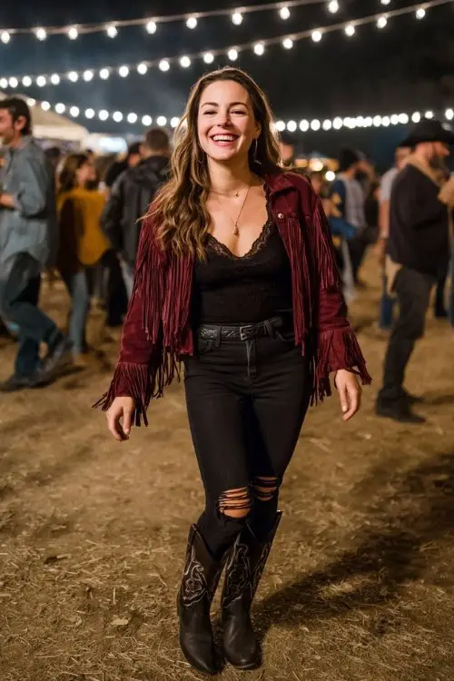 A curvy woman wears ripped black jeans with tall black cowboy boots and a burgundy fringe jacket
