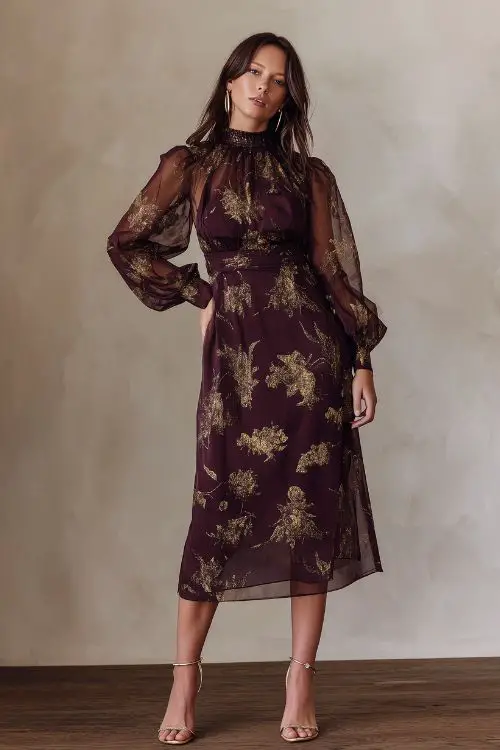 A full-body image of a woman in a deep plum midi dress with gold foil floral print, a high neckline