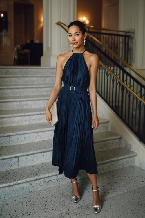 A petite woman wears a navy pleated halter midi with a keyhole back and thin belt
