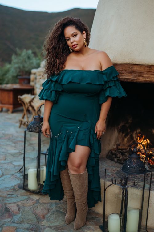 A plus-size woman over 40 wears a deep emerald off-the-shoulder gown with cascading ruffle details and subtle gold embroidery, paired with elegant suede knee-high boots