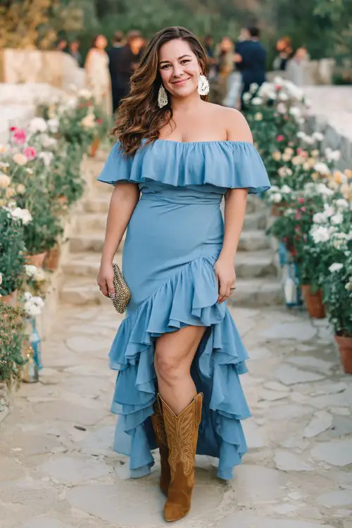 A plus size woman wearing a dusty blue off-shoulder dress with layered ruffles, suede cowboy boots, statement earrings