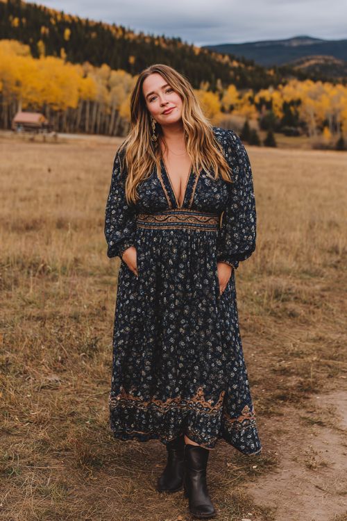 A plus-size woman wears a navy floral maxi dress with sheer puff sleeves and bronze embroidery, styled with dark leather ankle boots