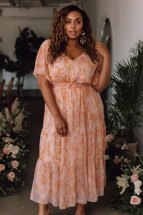 A plus-size woman wears a peach floral maxi dress featuring delicate floral prints, sheer short sleeves, and a relaxed tiered silhouette