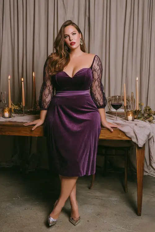A plus-size woman wears a plum velvet midi with illusion lace sleeves and a defined waist, styled with metallic block heels and drop earrings