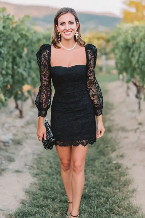 A woman over 30 wears a black lace mini dress featuring a structured square neckline, puff sleeves, and scalloped lace trim along the hem with subtle pleated underlayers