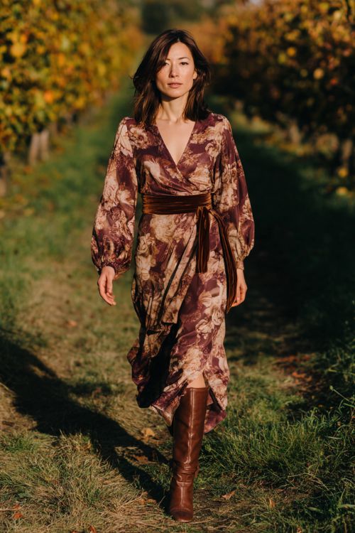 A woman over 30 wears a cinnamon floral wrap dress with dramatic balloon sleeves and velvet ribbon trim, styled with heeled knee-high boots