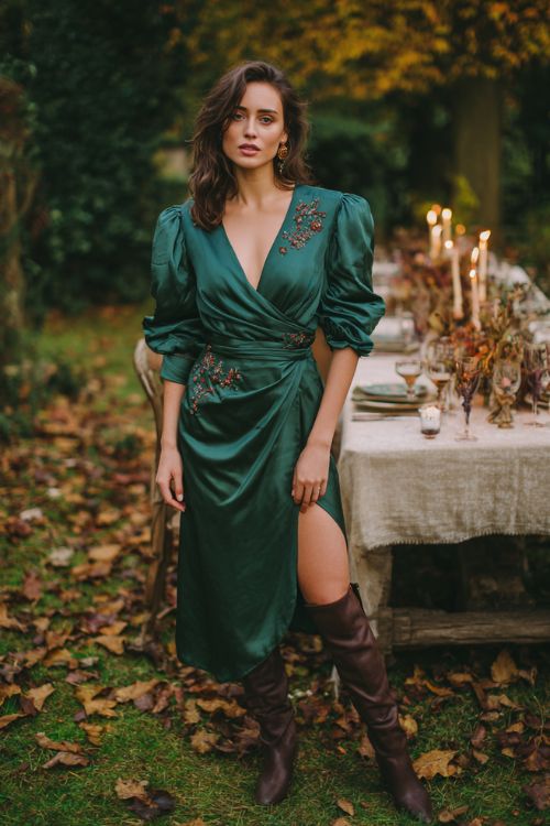 A woman over 30 wears a forest green satin wrap dress with dramatic puff sleeves and delicate floral appliqués, styled with chocolate brown knee-high boots