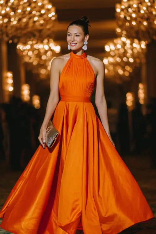 A woman over 30 wears a vivid sunset-orange halter neck dress featuring a pleated satin skirt and open-back design, styled with chandelier earrings