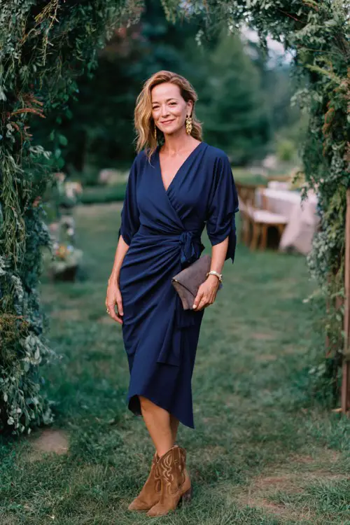 A woman over 40 wearing an elegant navy wrap dress with draped sleeves, suede cowboy boots, a structured clutch and statement earrings