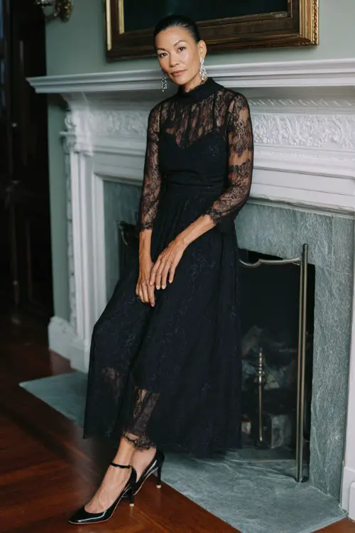 A woman over 40 wears a black lace midi dress featuring a high neckline, sheer lace sleeves, and layered chiffon panels