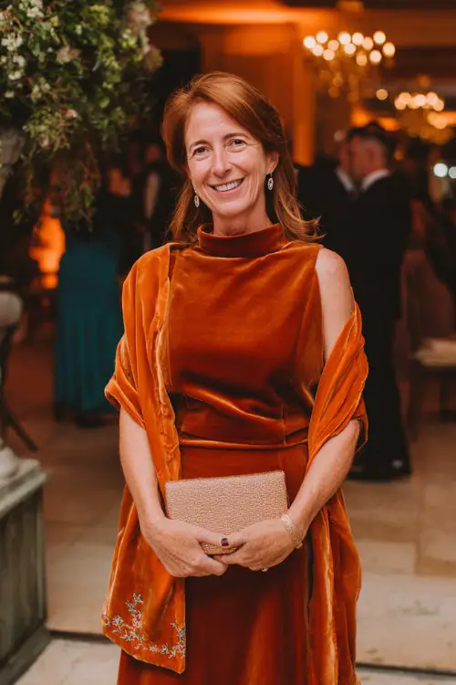 A woman over 40 wears a burnt-orange velvet midi dress featuring a high neckline, subtle embroidery along the sleeves, and a softly flared skirt