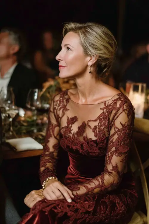 A woman over 40 wears a deep rust lace gown featuring sheer long sleeves, intricate floral patterns, and a graceful A-line skirt, accessorized with a velvet wrap and pearl jewelry