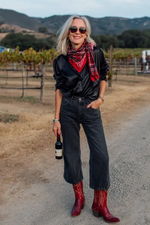A woman over 40 wears dark bootcut jeans with red snakeskin cowboy boots, styled with a black satin blouse and plaid scarf