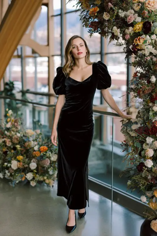A woman over 50 wears a black velvet midi dress with sheer puffed sleeves