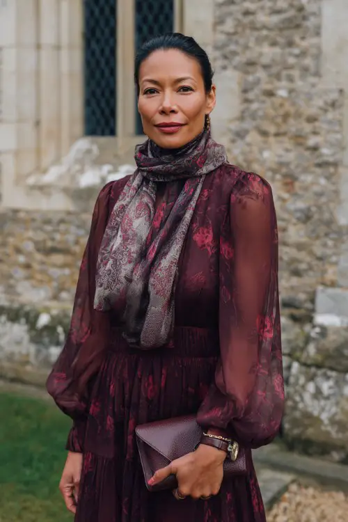 A woman over 50 wears a maroon floral-print dress with a sheer layered skirt and puff sleeves, wrapped in a silky patterned scarf