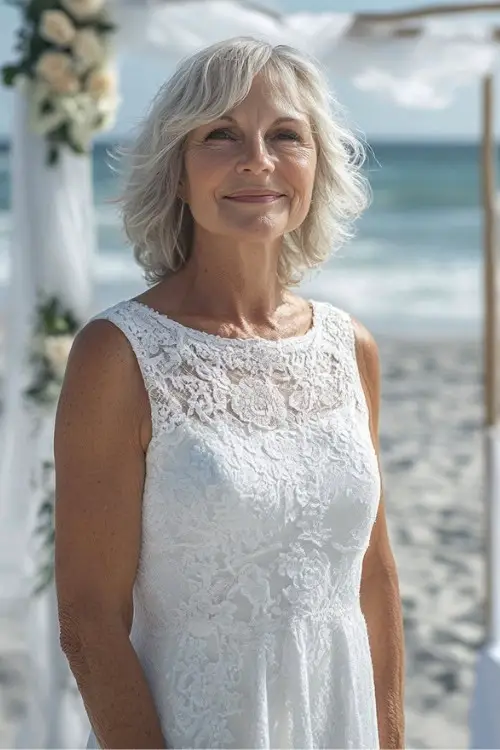 A woman over 50 wears a sleeveless white lace wedding guest dress at a beachside ceremony