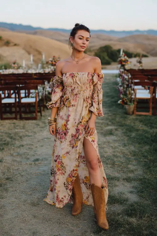 A woman wearing a bold floral off-shoulder maxi dress with a thigh slit, tan cowboy boots, a statement cuff bracelet