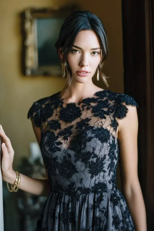 A woman wears a black lace dress featuring delicate floral appliqué over a soft neutral-toned lining, cap sleeves, and a gently pleated skirt