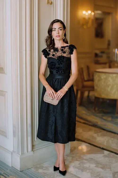 A woman wears a black lace dress featuring delicate floral appliqué over a soft neutral-toned lining, cap sleeves