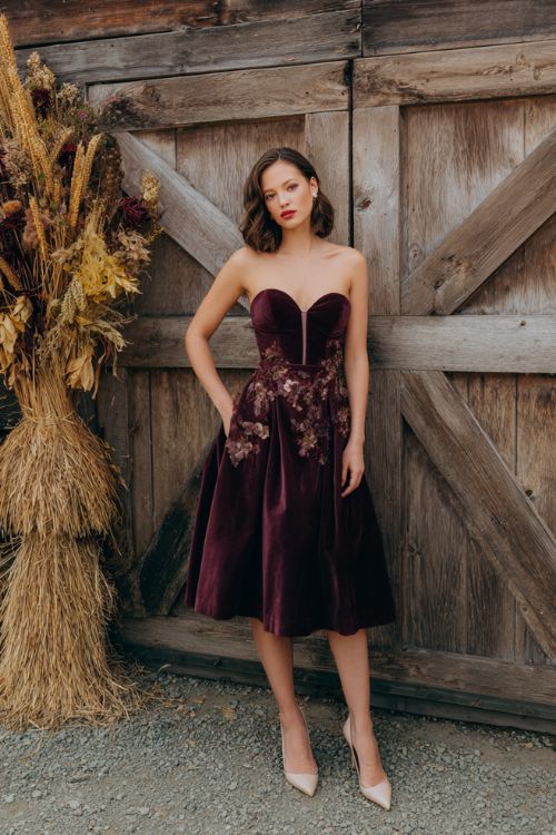 A woman wears a plum-colored fit-and-flare dress with velvet floral appliqués and a sweetheart neckline, paired with pointed-toe pumps
