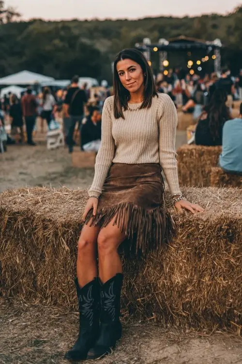 A woman wears a suede fringe skirt with a fitted knit sweater and black cowboy boots