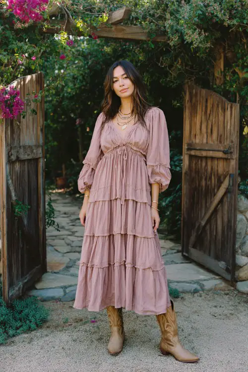 A woman wears a tiered chiffon dress in dusty rose with airy layers, paired with light brown cowboy boots and delicate jewelry