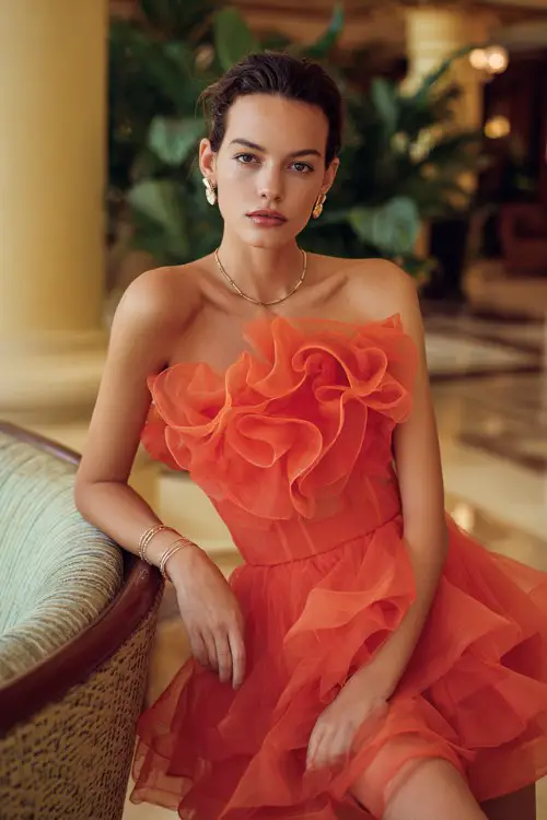 A woman wears a vibrant coral-orange organza dress with sculptural ruffles across the bodice and a dramatic layered skirt