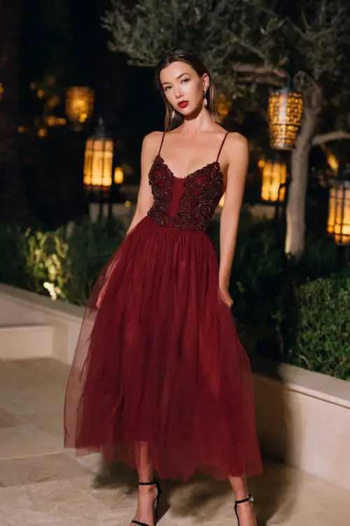 A woman wears a wine-colored gown with a tulle skirt and floral embroidered bodice, styled with black strappy heels