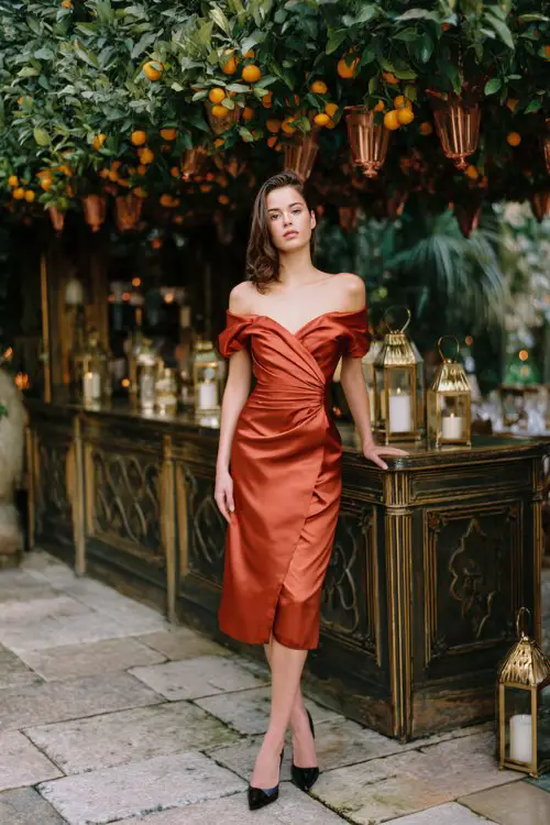 A woman wears an rust satin midi with an off-shoulder neckline, wrap waist, and tulip hem