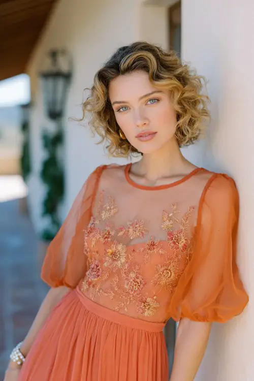 Short tangerine orange chiffon dress with sheer puff sleeves, intricate floral embroidery across bodice and sleeves, flowy layered skirt