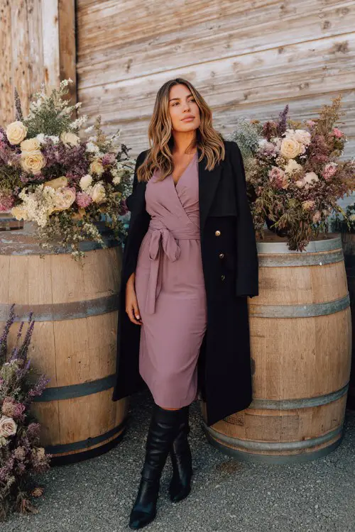 A curvy woman wears a mauve midi dress with a belted black coat and knee-high boots