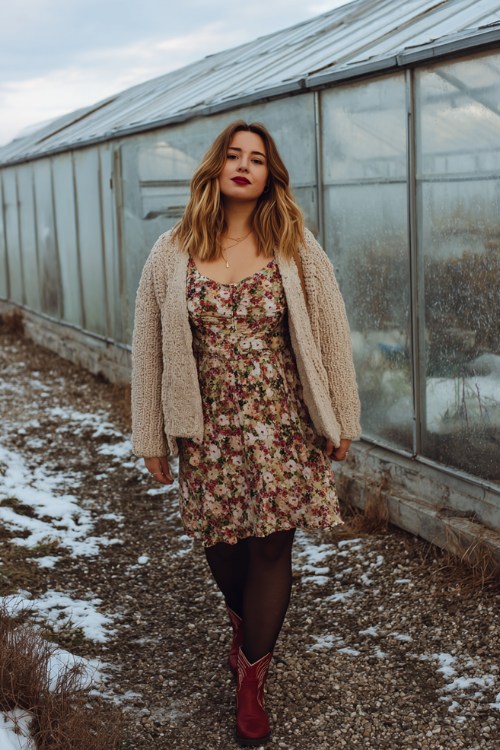 A curvy woman wears a tiered floral midi dress with black tights, an oversized cardigan, and red cowboy boots