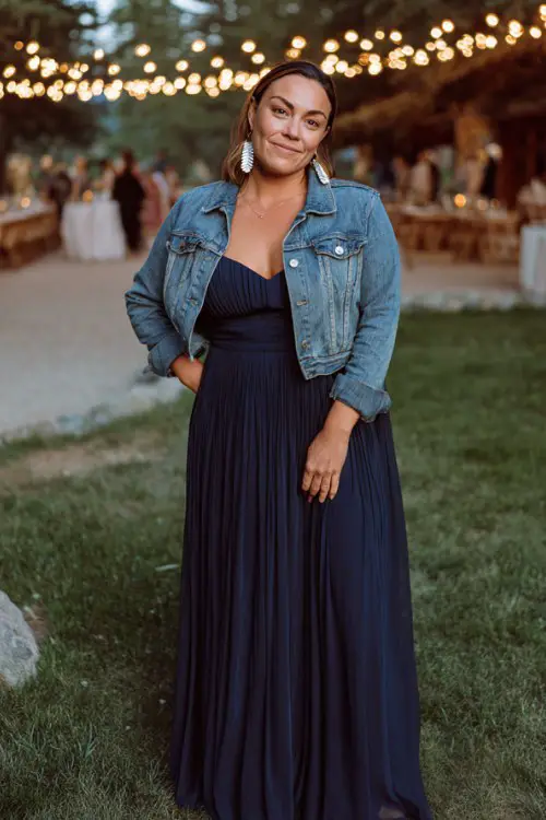 A full body image of woman over 30 wears a navy chiffon maxi dress with pleated details, styled with a cropped denim jacket and silver earrings