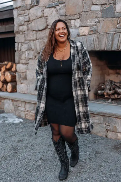 A plus-size woman wears a black bodycon dress layered under a long plaid coat, paired with thick tights and tall black cowboy boots