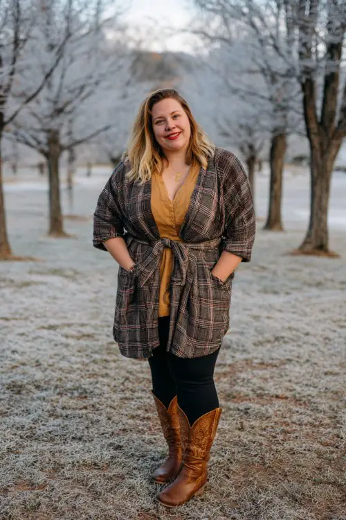 A plus-size woman wears high-rise black leggings with brown cowboy boots, a knit tunic, and a plaid wrap coat